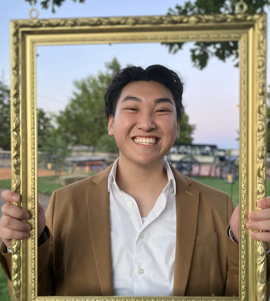 Darren Don smiling, holding up an empty gold picture frame around himself outdoors at dusk.